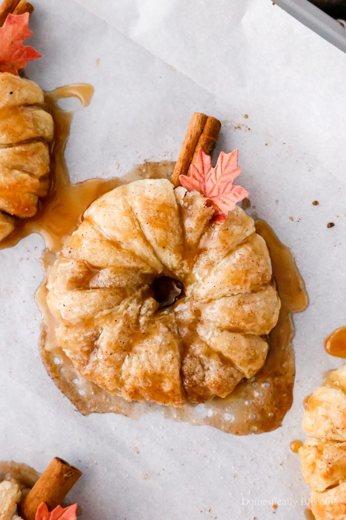 These Apple Cinnamon Pastry Donuts are filled with fresh apple slices, surrounded by puff pastry, covered in cinnamon sugar, drizzled with caramel sauce, and topped with a cinnamon stick and edible maple leaf.