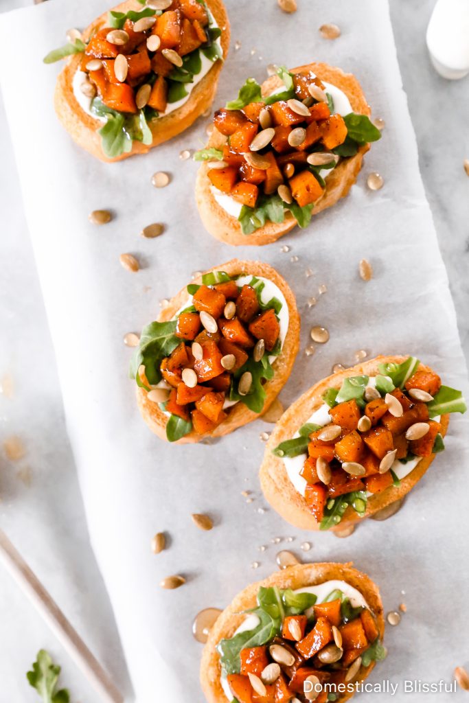 This Whipped Goat Cheese Butternut Squash Bruschetta begins with sliced toasted baguette, topped with whipped goat cheese, arugula, roasted butternut squash, honey, and pumpkin seeds.  
