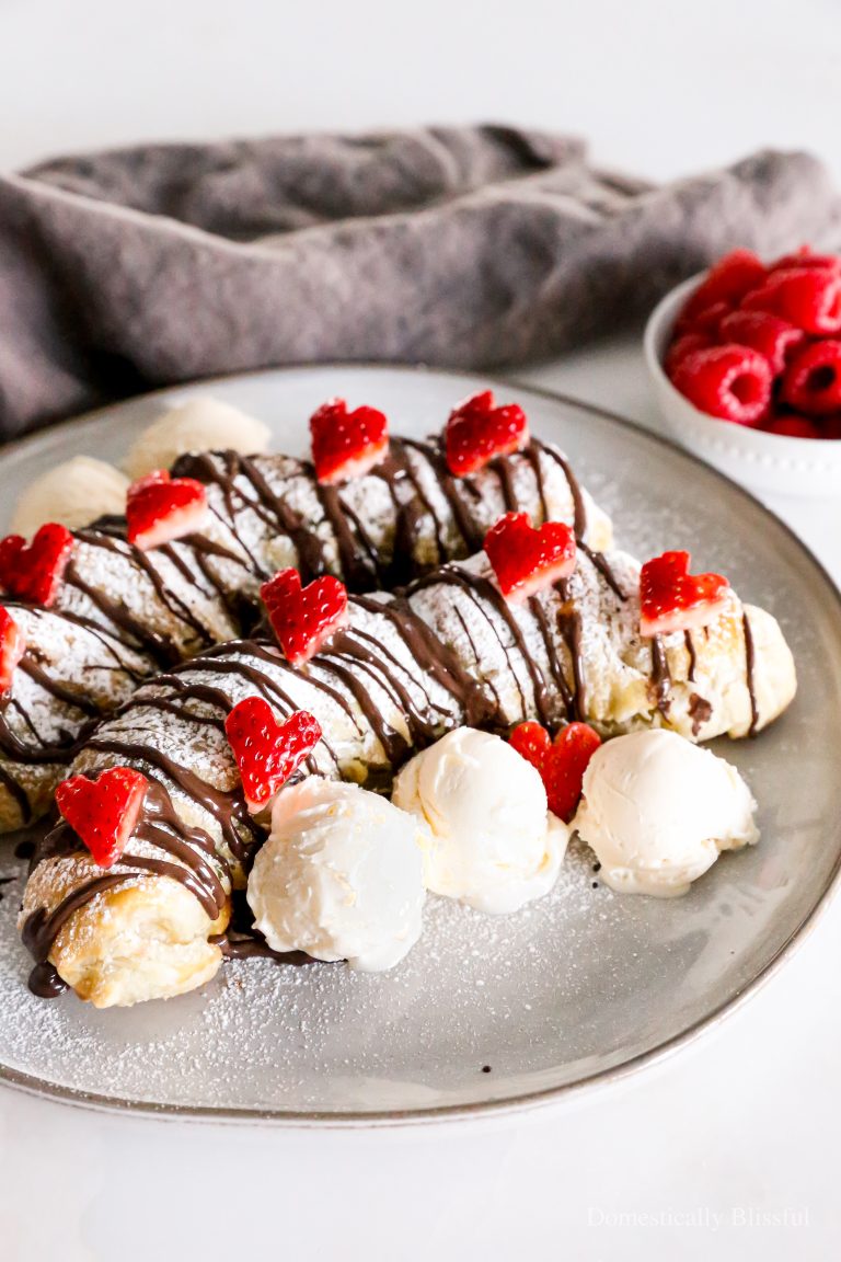 This Puff Pastry Banana Split is filled with Nutella and raspberries, wrapped in puff pastry, and topped with powdered sugar, dark chocolate drizzle, ice cream, and heart strawberries. Perfect for Valentine's Day or a Galentine's party!