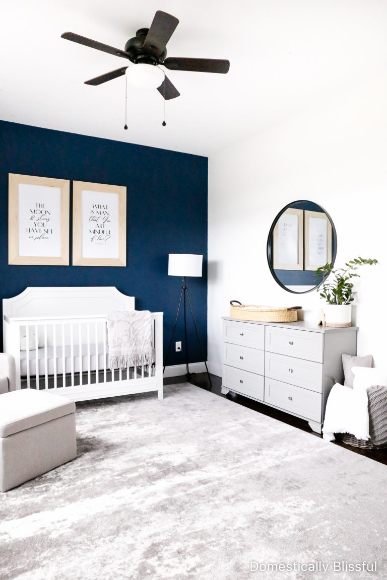 A simple Space Themed Nursery with a dark blue wall, outer space printables, grey glider, white crib, and grey dresser topped with a baby changing basket.