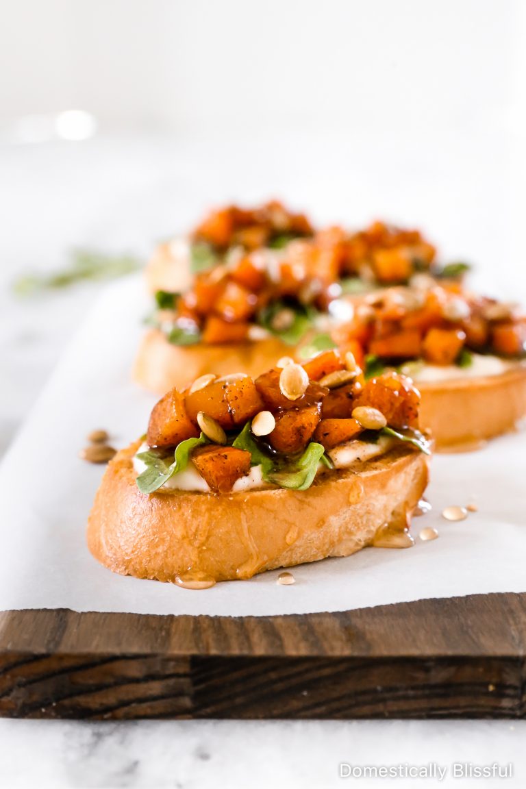 This Whipped Goat Cheese Butternut Squash Bruschetta begins with sliced toasted baguette, topped with whipped goat cheese, arugula, roasted butternut squash, honey, and pumpkin seeds.